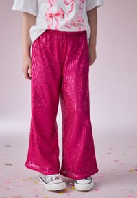 Pantaloni con sovrapposizione di paillettes fucsia, design a gamba larga, superficie testurizzata; abbinati a sneaker bianche. Elementi decorativi sulla camicia sopra.
