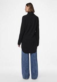 Schwarzer übergroßer Pullover mit hohem Kragen, gerippter Struktur und geschwungenem Saum. Kombiniert mit weitem, hellblauem Jeansstoff.