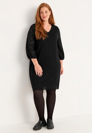 Schwarzes Spitzenkleid mit Dreiviertelärmeln, V-Ausschnitt und gewelltem Saum, getragen über schwarzen Strumpfhosen mit schwarzen Slippers.