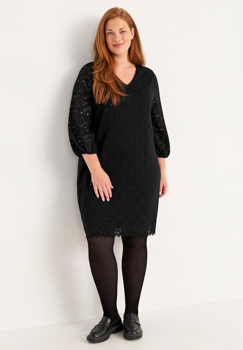 Svart spetsklänning med trekvartsärmar, V-ringning och scalloped fåll, bärs över svarta strumpbyxor med svarta slip-on-skor.