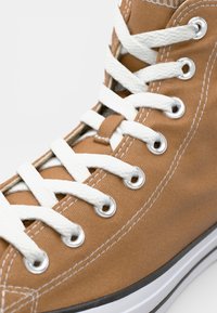 Hoge canvas sneaker in tan met witte oogjes en veters. Heeft een platte zool en een contrasterende zwarte streep langs de onderkant.