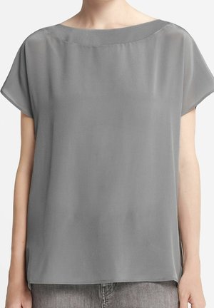 Femme portant un chemisier gris ample à manches courtes avec un col bateau, associé à un jean en denim gris clair.