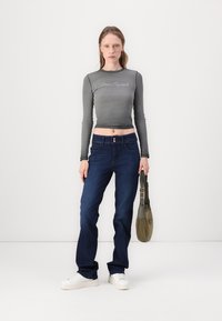 Grå langermet cropped genser med logo, kombinert med mørkeblå høytaljede jeans og hvite sneakers. En grønn veske henger fra den ene skulderen.