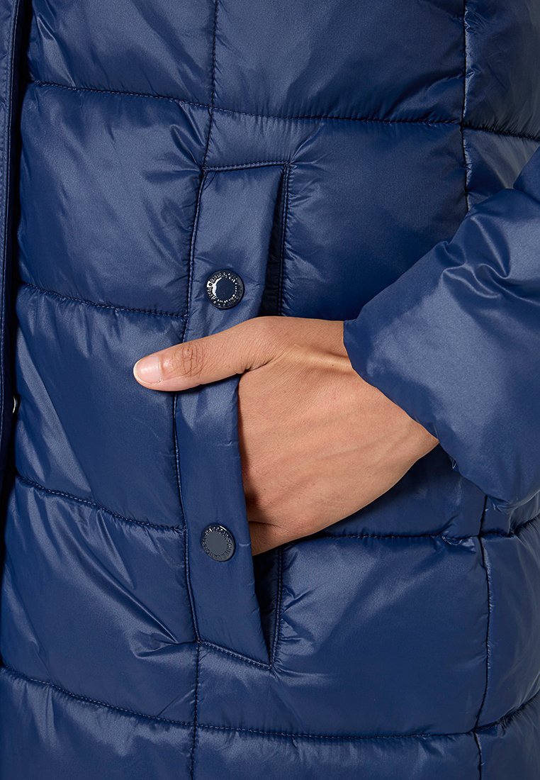 Mano inserita nella tasca laterale di un giubbotto imbottito blu navy con chiusure a bottoni a pressione.