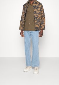 Kurtka pikowana w kamuflażowym wzorze w odcieniu brązu, noszona na oliwkowej koszuli z guzikami, w połączeniu z jasnoniebieskimi dżinsami w kształcie dzwonów i kremowymi trampkami.