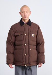 Chaqueta acolchada marrón con un cuello de pana negro, herrajes dorados, dos bolsillos en el pecho y cordón ajustable en el dobladillo.