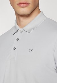 Svijetlosiva polo majica s ovratnikom, dvobuttonim kopčanjem i CK logotipom izvezanim u tamnosivoj boji na prsima.