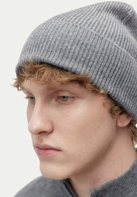 Grå, ribbad mössa tillverkad av mjukt stickat material, sitter tajt och har en strukturerad yta. Närbild på ett ansikte med ljus hy och något rufsigt hår.