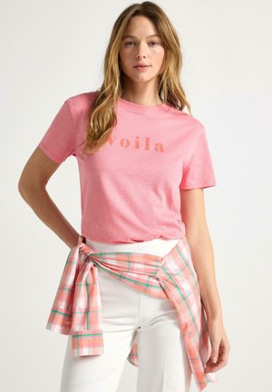 Kvinde iført en lyserød "voila" t-shirt, hvide bukser og en lyserød ternet skjorte bundet om livet, stående foran en ensfarvet baggrund.