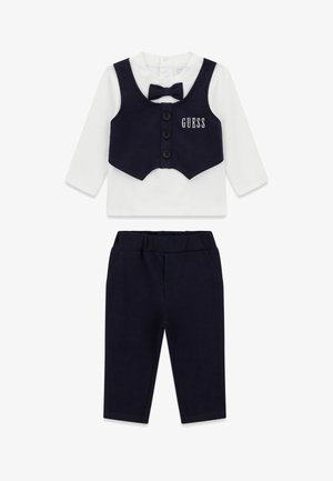 Tenue habillée pour bébé comprenant un haut blanc à manches longues, un gilet bleu marine avec nœud papillon, et un pantalon assorti bleu marine, avec le logo "GUESS" sur le gilet.