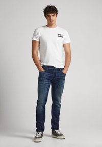Camiseta blanca de manga corta con el logo negro de "Pepe Jeans", combinada con jeans de mezclilla azules y zapatillas grises con suelas blancas.