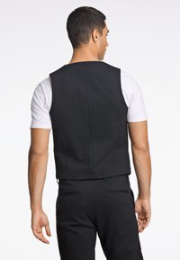 Schwarze ärmellose Weste mit glatter Textur, klaren Linien und geradem Schnitt, getragen über einem weißen Hemd. Keine sichtbaren Beschläge oder Verzierungen.