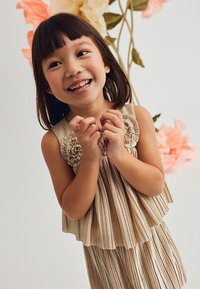 Abito beige senza maniche con design plissettato, caratterizzato da accenti floreali sulle spalle. Tessuto liscio e lucido con un aspetto a strati.