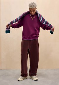 Fiatal személy rövid szőke hajjal, lila pulóvert, barna laza nadrágot és sportcipőt visel, és egy csíkos sálat tart a nyaka mögött.