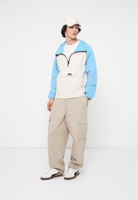 Leichter, zweifarbiger Pullover mit Reißverschluss in Beige und Himmelblau, mit einer Fronttasche, kombiniert mit beigen Cargo-Hosen und weißen Sneakers.