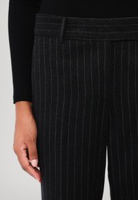 Pantalons en laine noirs avec des rayures grises, présentant une coupe ajustée et une taille plate, montrés avec une main partiellement visible reposant sur le tissu.