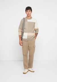 Νεαρός άνδρας στέκεται φορώντας πλεκτό πουλόβερ σε μπεζ, άσπρο και γκρι χρώματα, μπεζ cargo παντελόνι, άσπρα αθλητικά παπούτσια και γκρι τσάντα ώμου σε απλή λευκή φόντο.