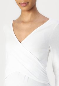 Witte langwerpige top met een v-hals en wikkelontwerp. Zachte stof met een gladde textuur; heeft plooien bij de taille voor een flatterende pasvorm.