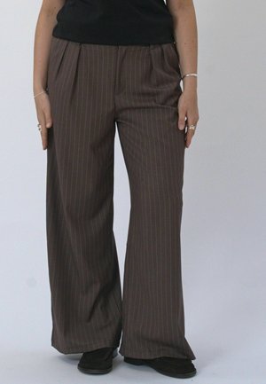 Persoon die bruine pinstripe wijde broek, zwarte schoenen en een zwarte top draagt, staand tegen een effen lichte achtergrond.