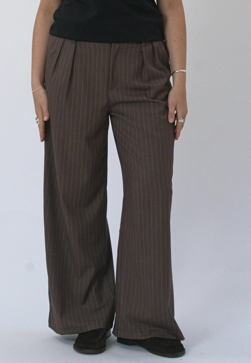 Personne portant un pantalon large marron à fines rayures, des chaussures noires et un haut noir, debout devant un fond clair uni.
