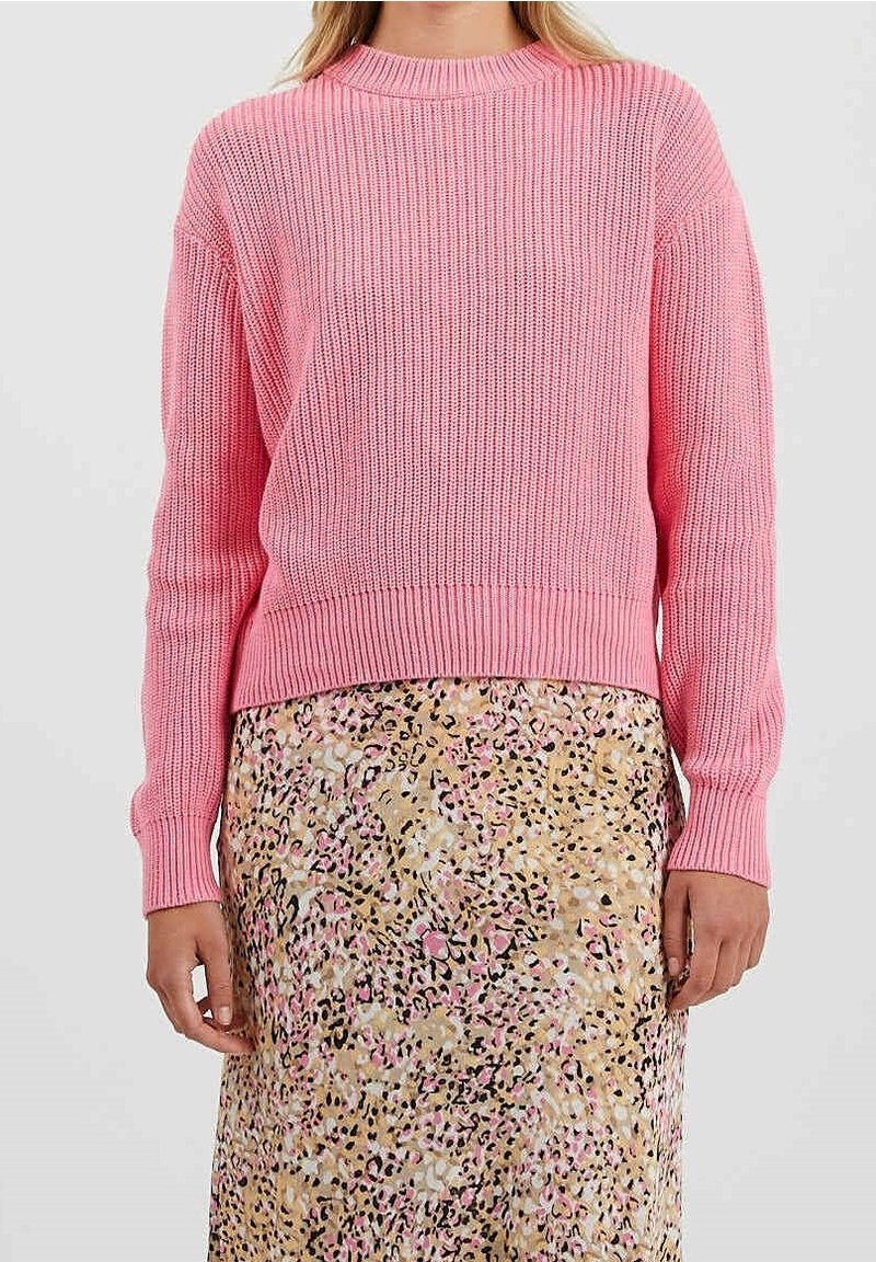 Personne portant un pull en tricot rose et une jupe beige, rose et noire à motifs, debout devant un fond clair uni.