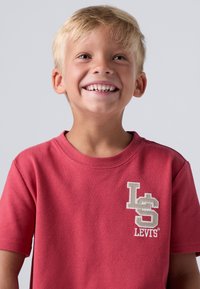 Raudona medvilninė marškinėliai su trumpomis rankovėmis, su siuvinėtu "L" ir "S" logotipu smėlio spalvos kairėje krūtinės dalyje.