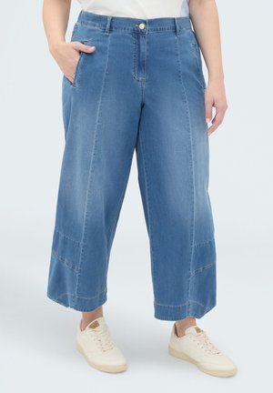 Osoba nosząca niebieskie na środku szerokie, krótkie dżinsy z przednimi szwami i beżowe trampki, stojąca z jedną ręką w kieszeni.