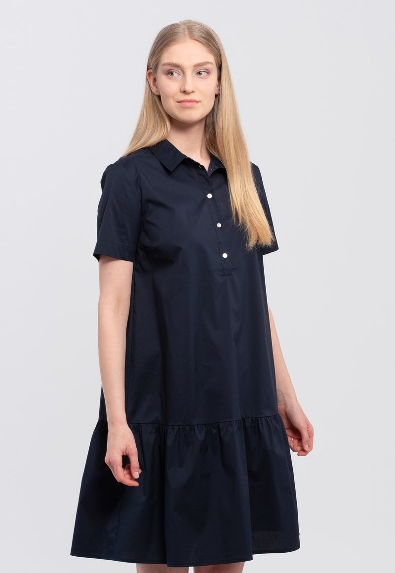 Navyblauwe jurk van katoen; beschikt over een knoopsluiting bij de kraag, korte mouwen en een geplooide ruches-zoom. Glad textuur door de gehele jurk.