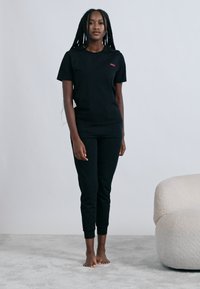 Fekete pamut rövid ujjú, karcsúsított fazonú póló, fekete karcsúsított nadrággal párosítva. A pólón a mellrészen egy kisméretű piros logó található.