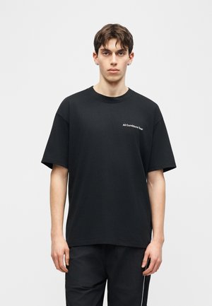 Mladý muž s krátkymi hnedými vlasmi stojí čelom vpred, oblečený v čiernom oversized tričku s nápisom „All Conditions Gear“ na hrudi a v čiernych nohaviciach.