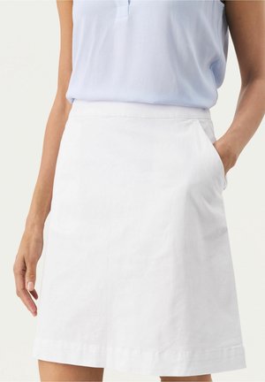 Kvinde iført en lyseblå ærmeløs top stoppet ned i et hvidt knælængde skørt med højre hånd i lommen.