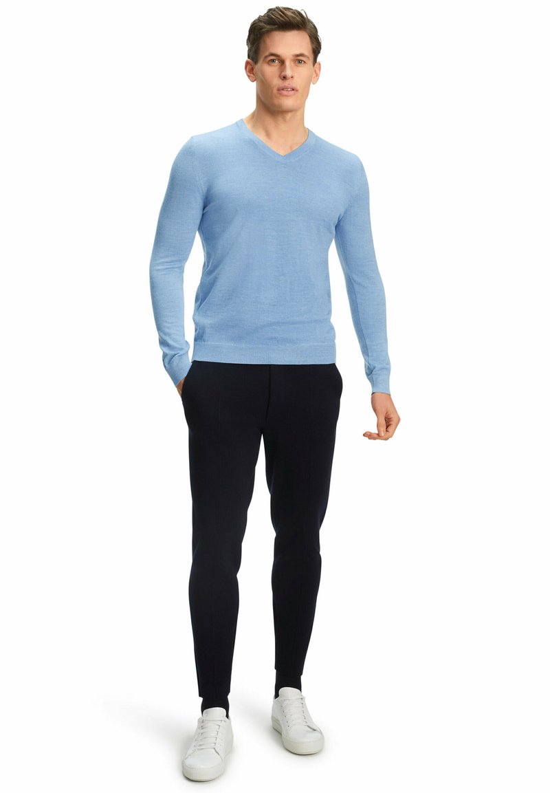 Hellblaues, tailliertes V-Ausschnitt-Pullover aus weichem Stoff, kombiniert mit schwarzen Slim-Fit-Hosen und weißen Turnschuhen, in stehender Pose. Einfaches Design.