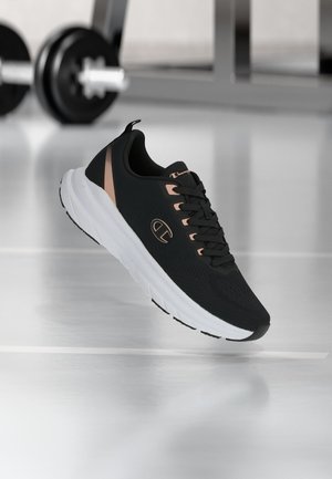 Schwarzer und beiger Champion-Laufschuh mit weißer Sohle, in der Luft positioniert in einem Fitnessstudio mit verschwommenem Gewichtheber-Equipment im Hintergrund.