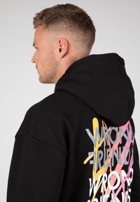 Felpa con cappuccio nera con una grafica sul retro che presenta un design di testo sovrapposto e colorato in rosa, giallo e grigio. Tessuto morbido, vestibilità casual.