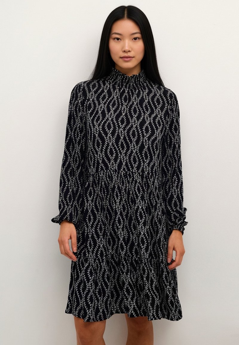 Zwarte jurk met een hoge hals, lange mouwen en een vloeiende silhouet met een witte golvende patroon. Zacht stof met een textuurrijke uitstraling.