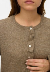 Sieviete valkā brūnu adītu kardiganu ar volānu detaļu un baltām pogām, roka atbalstīta uz apakšējās pogas zonas.