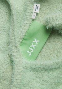 Lichtgroene fuzzy trui stof met een gestructureerd oppervlak. Bevat een label met "JUXX" in het wit, een label dat maat "S" aangeeft, en verzorginstructies.