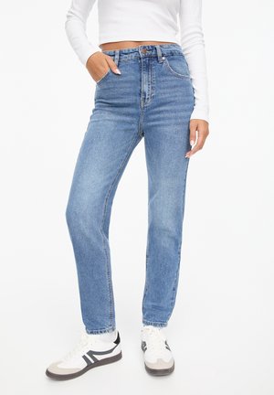 Femme portant un jean bleu clair taille haute à jambes droites, des baskets blanches et un top blanc court à manches longues, main dans la poche.