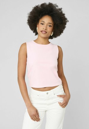 Femme avec une coiffure afro naturelle portant un haut rose clair sans manches et un pantalon blanc, debout avec une main dans la poche sur un fond uni.