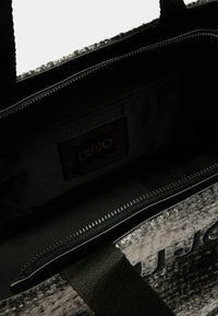 Sort tote taske med tekstureret materiale og en lynlås lomme indeni. Har et fremtrædende LIU JO-logo på en glat baggrund.