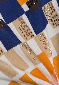 Gros plan sur un tissu avec un motif en grille coloré comprenant des teintes bleues, orange, beige et blanches, décoré de clous métalliques rectangulaires brillants.