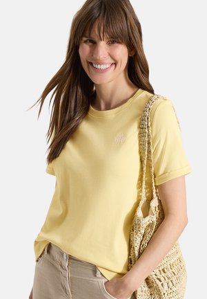 Uśmiechnięta kobieta z długimi brązowymi włosami, ubrana w żółtą koszulkę i beżowe spodnie, niosąca plecioną beżową torebkę na ramię.