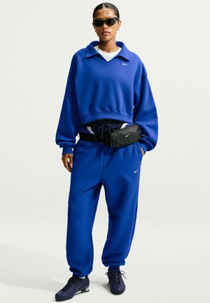 Femme portant un survêtement bleu Nike, un sac banane noir, des lunettes de soleil foncées et des baskets noires, debout avec une main dans la poche.