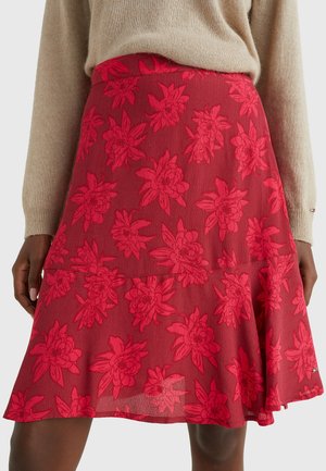 Falda roja con estampado floral en línea A y dobladillo en capas, combinada con un suéter beige de manga larga, mostrada desde la cintura hasta la mitad del muslo en una persona de piel oscura.
