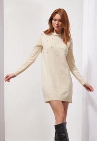 Robe en tricot de couleur crème, à manches longues, avec un col rond et des embellissements. Présente une coupe décontractée et des poignets côtelés, arrivant mi-cuisse.