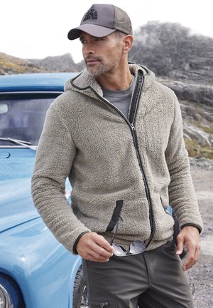 Fleece zip-up hoodie in cream with a textured exterior, black zipper, and two side pockets. Worn with a gray t-shirt and glasses in hand.