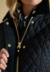 Jachetă matlasată de culoare bleumarin cu fermoar auriu, butoane cu capse și guler din catifea. Strat interior cu dungi vizibil. Țesătură texturată cu model în diamante.