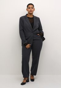 Costum gri croitorit cu un închidere cu un singur buton, rever structurat și pantaloni asortați. Sub acesta, un top din plasă cu buline negre.