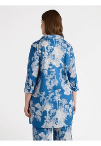 Niebieska odzież z rękawami do łokcia, z białym tropikalnym wzorem, akcentami guzików oraz luźnym krojem. Materiał wydaje się gładki i lekki.