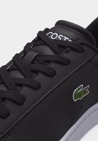 Μαύρο δερμάτινο αθλητικό παπούτσι με μαύρα κορδόνια, λευκή σόλα, την επιγραφή Lacoste στη γλώσσα και πράσινο λογότυπο κροκοδείλου στο πλάι.